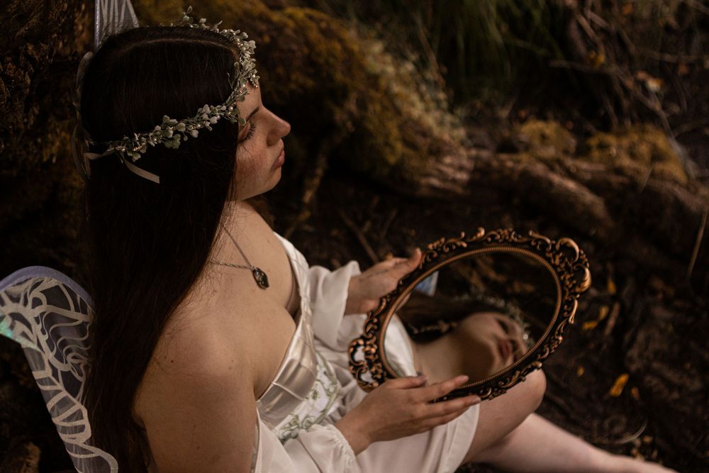 Young model dressed in white as a fairy holding a small vintage looking mirror in the woods near Eugene