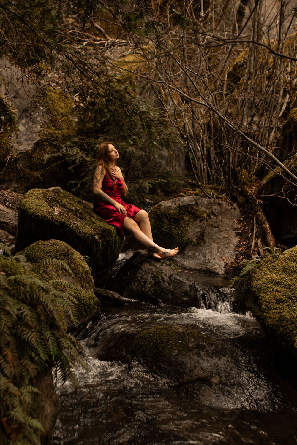 A client sits at the edge of the river, on some mossy rocks, wearing a red satin dress.