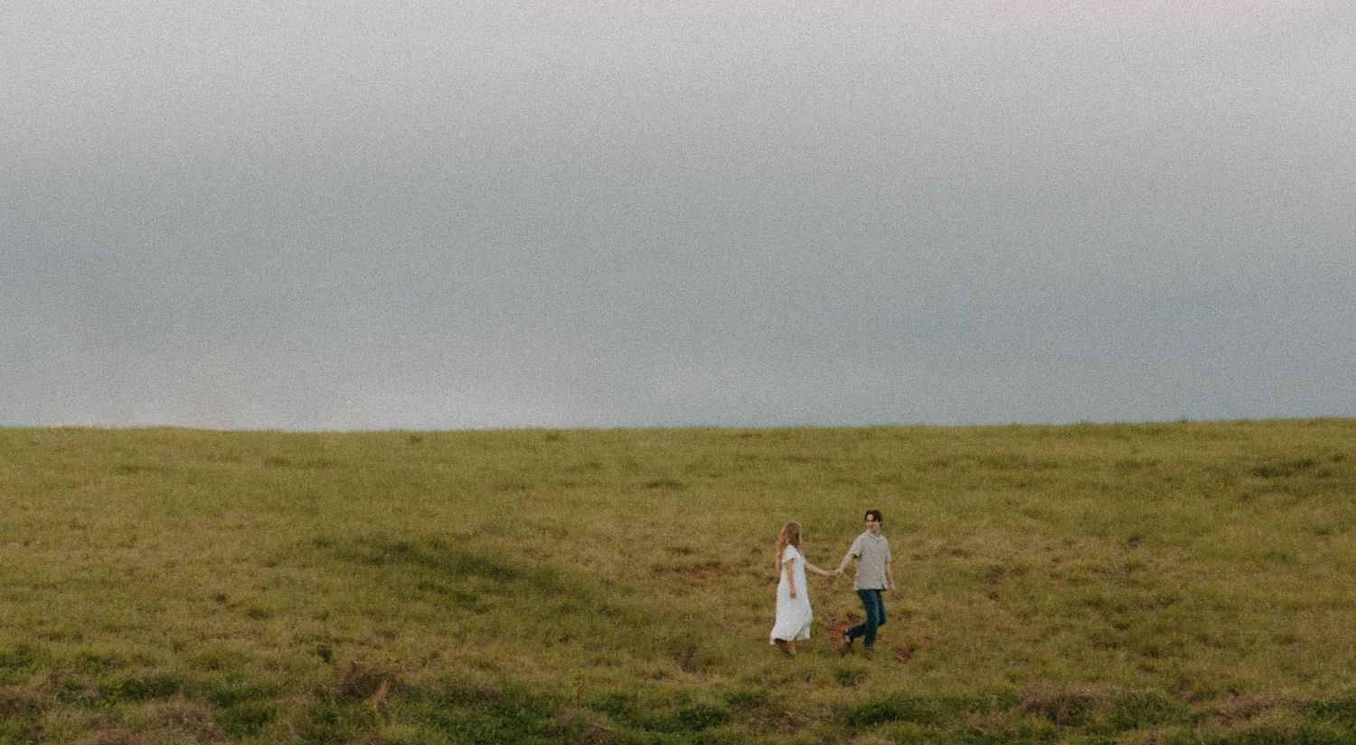 Two people walking together on a vast grassy hilltop against an overcast sky.