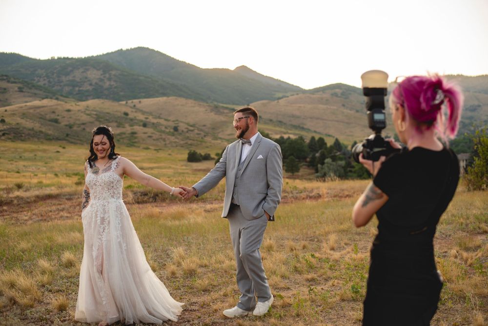 Photographer with vibrant hair captures couple posing in a scenic mountain meadow at sunset at The Manor House in Littleton, CO.