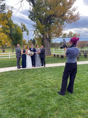 A photographer captures a wedding ceremony on a grassy hillside with autumn trees and white fencing in the background.
