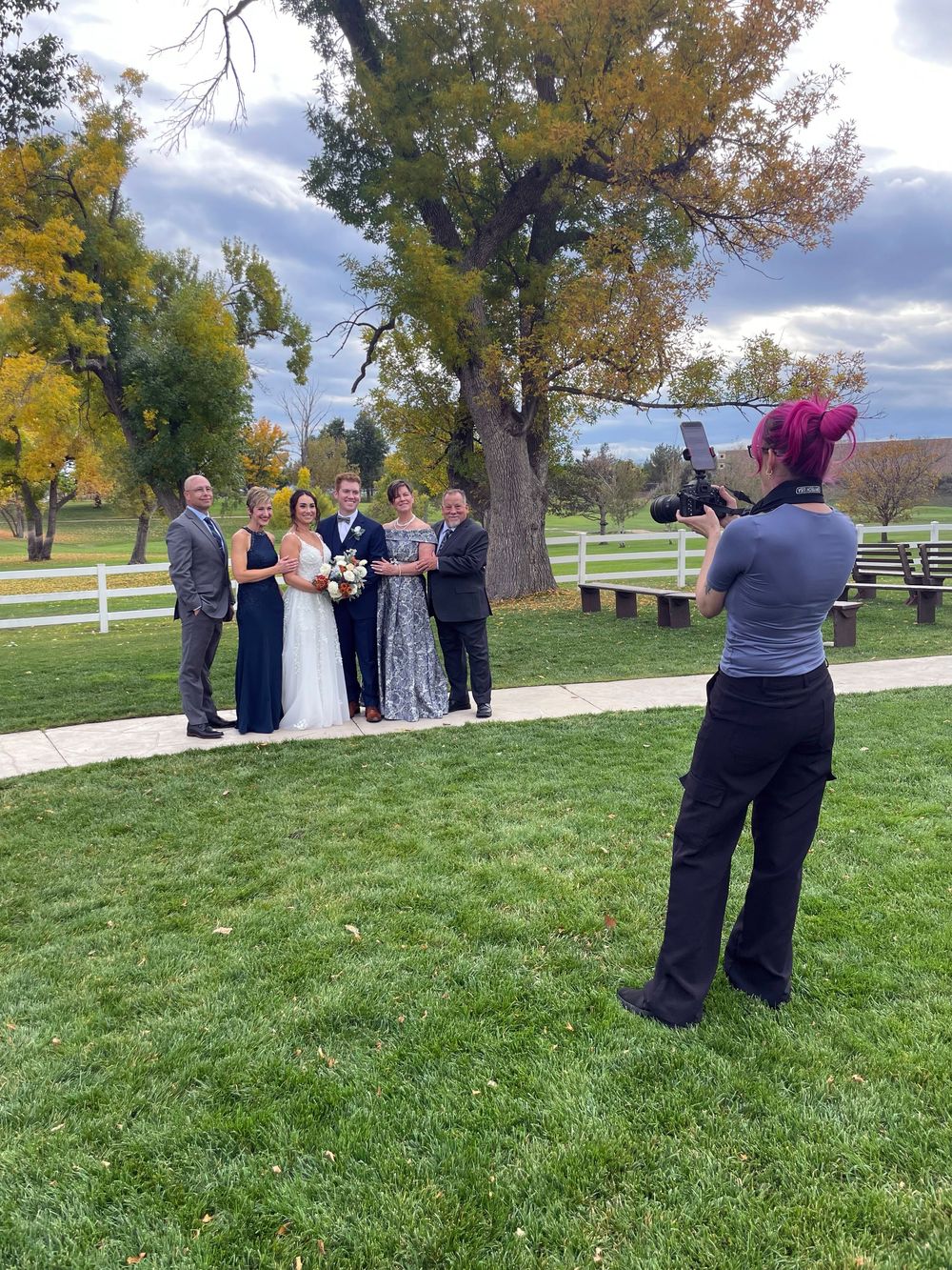 A photographer captures a wedding ceremony on a grassy hillside with autumn trees and white fencing in the background.