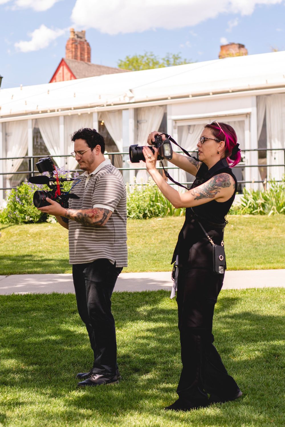 Two photographers capturing outdoor event on sunny day, woman with pink hair holding DSLR, man operating video camera on lawn.