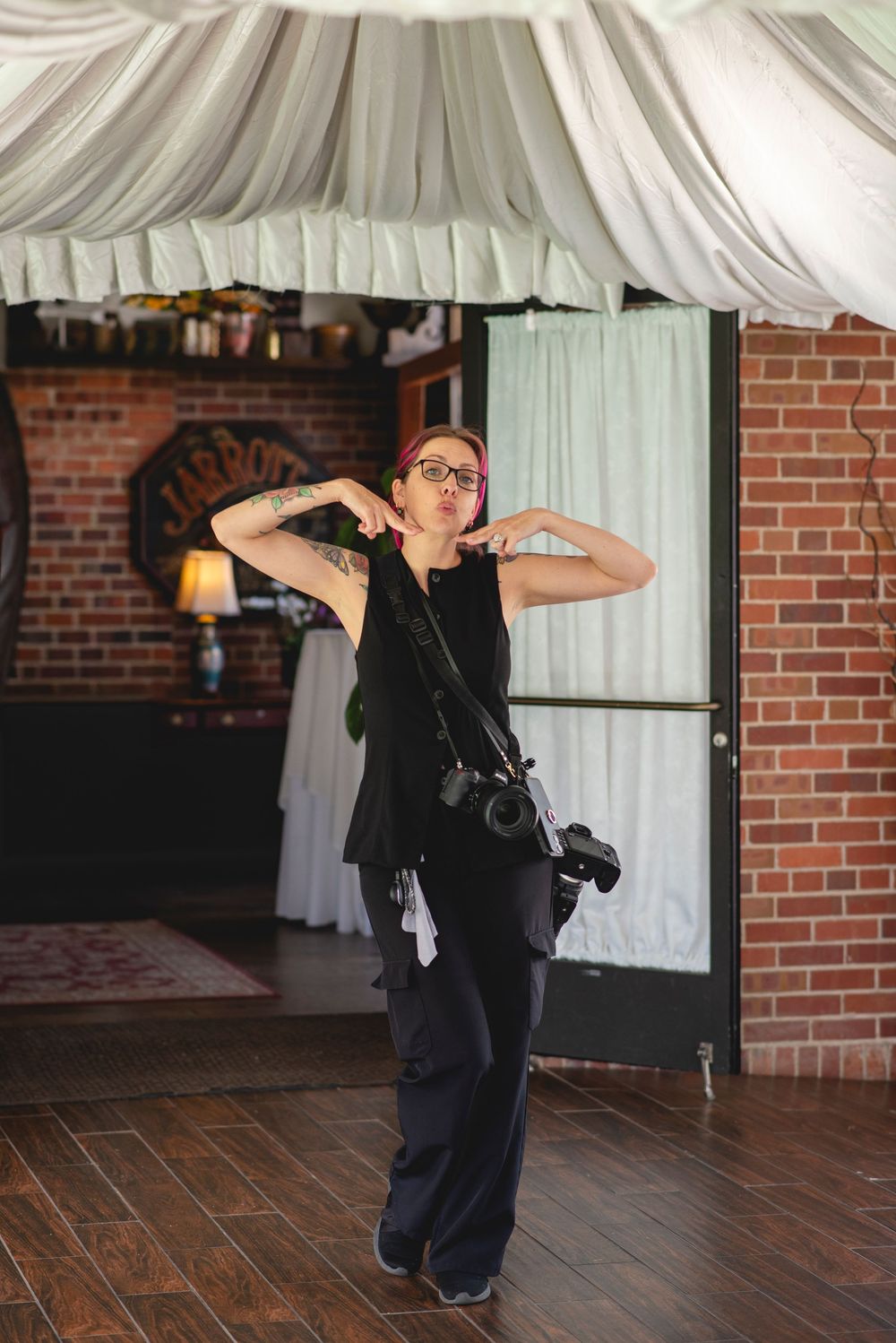 A tattooed female photographer in black outfit poses confidently indoors, camera on strap, brick wall backdrop.