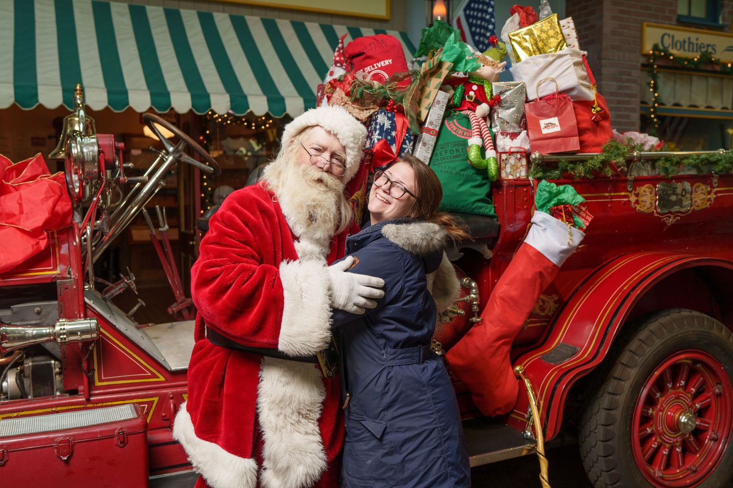 A festive holiday scene shows traditional Christmas figures beside a vintage red car filled with wrapped presents and decorations.