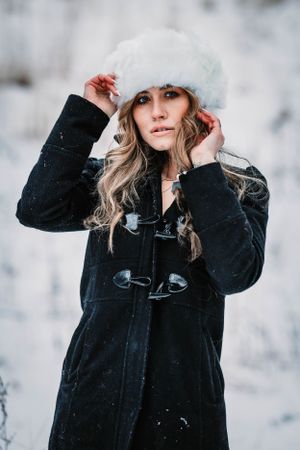Winter portrait in black toggle coat and white fur hat with falling snow.