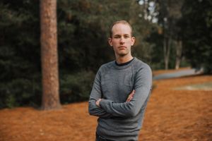 Person wearing grey sweater poses with crossed arms against autumn forest background.