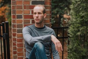 Person in grey sweater sits casually against brick pillar in outdoor setting.
