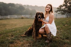 A brown dog poses with someone in a white dress in a pastoral farm setting at sunset.