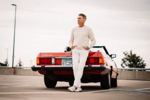 Red vintage Mercedes-Benz convertible parked in urban setting with driver in all-white attire.