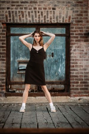 Model posing in black dress and sneakers against vintage blue door and brick wall background.