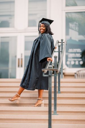 Graduate poses joyfully on concrete steps in traditional black academic regalia.