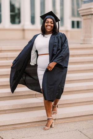 Graduate in cap and gown smiles while descending stone steps in elegant white dress.