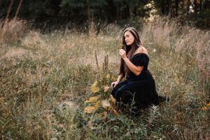 Woman in black dress kneeling thoughtfully in wildflower meadow.