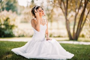 Bride in flowing white gown sitting gracefully on grass in sunlit garden.