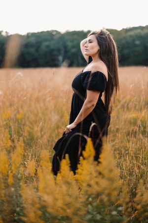 Woman in black dress poses dramatically in golden field at sunset.