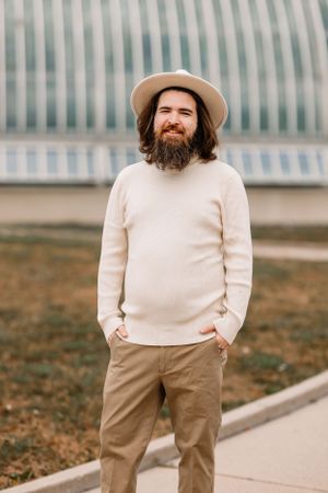 Bearded person in cream sweater and tan hat stands outdoors near modern architecture.