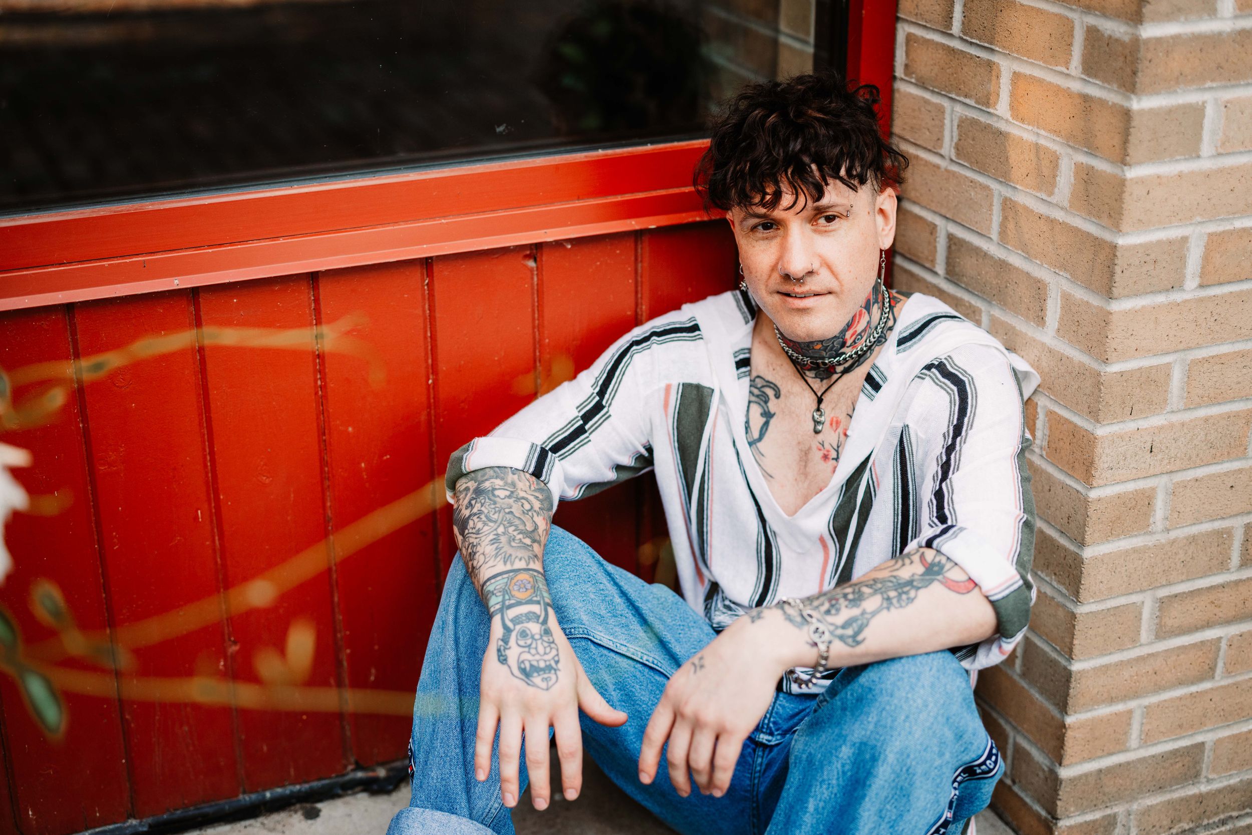 Casually dressed person with curly hair and tattoos sits against brick wall next to red doorway.
