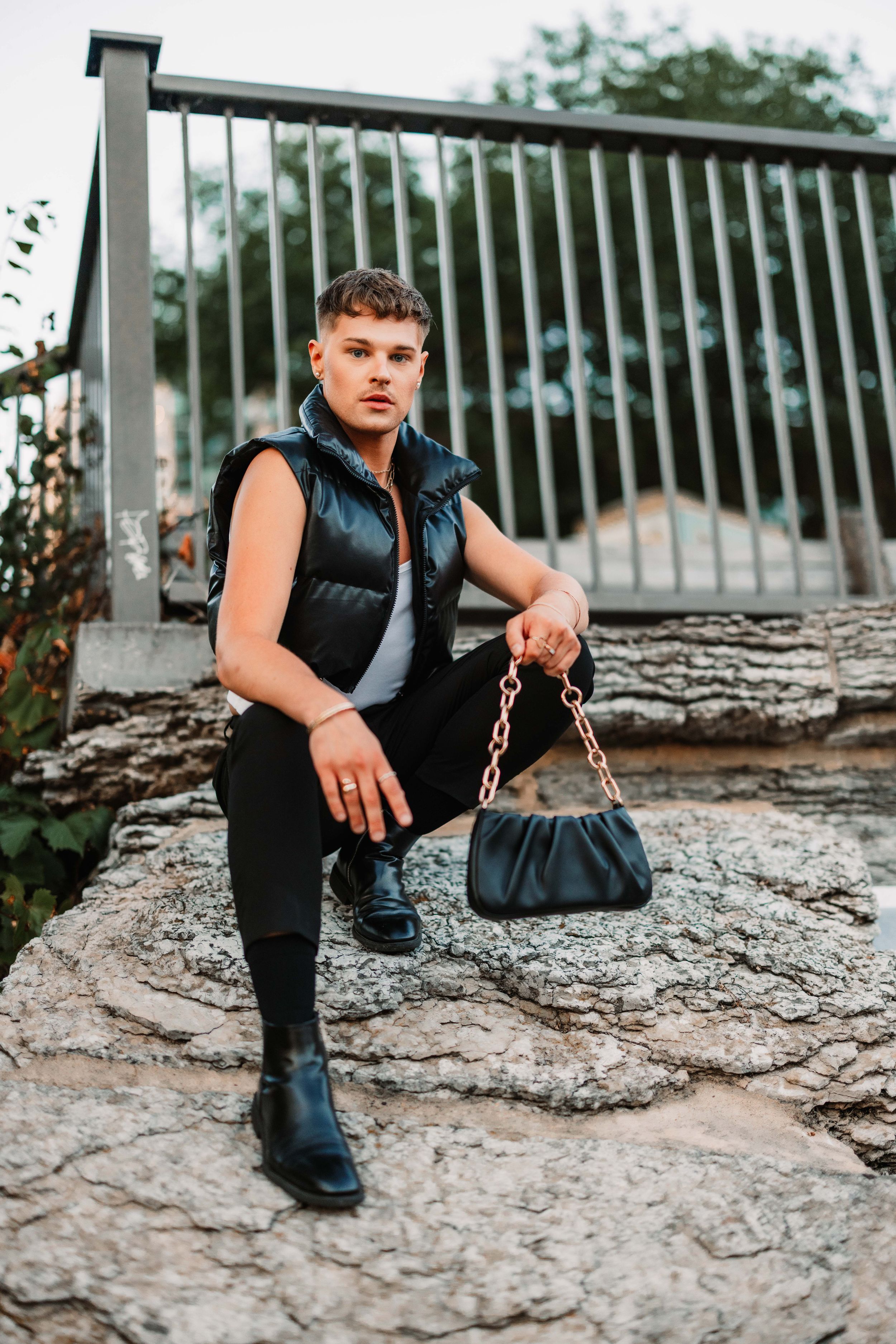 Person wearing black boots and vest sits on rocky ground while holding a dark handbag.