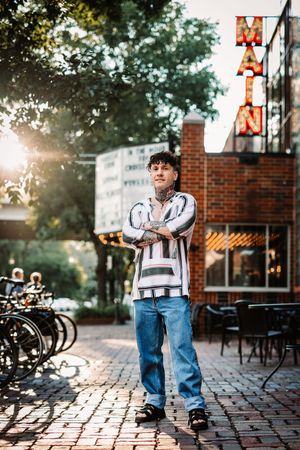 Person in denim jacket and jeans poses playfully on brick patio with sunlight streaming through trees during outdoor photoshoot.