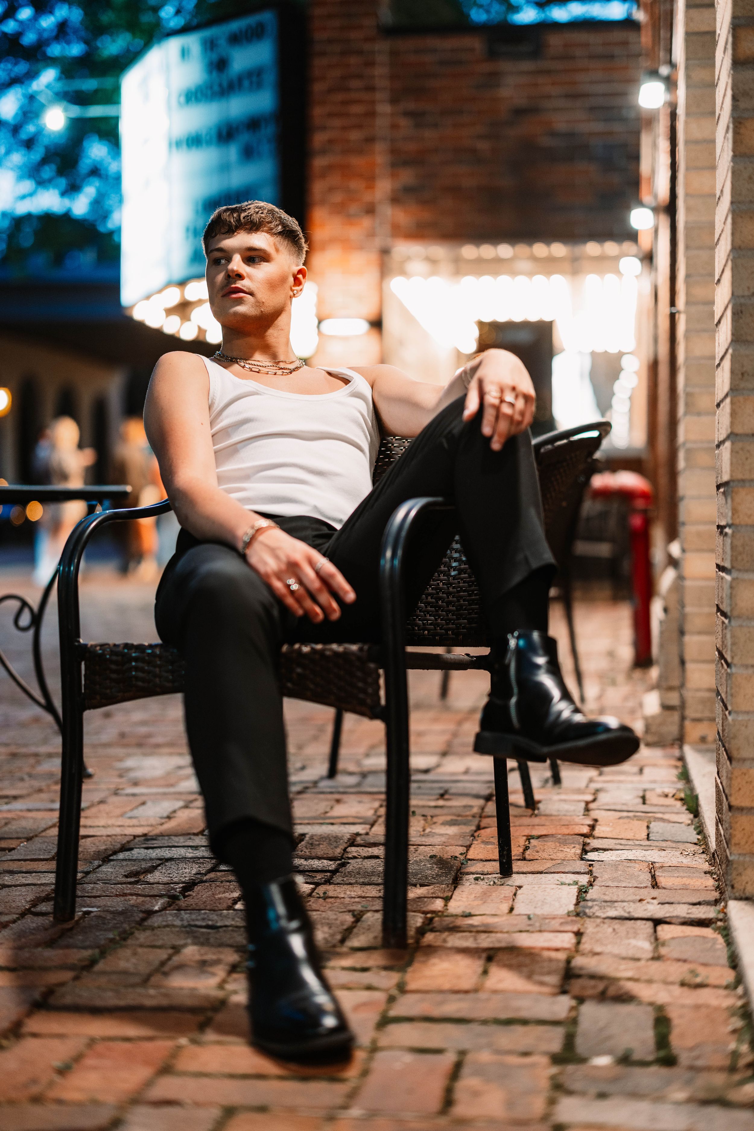 Person in black pants and boots sits in woven patio chair against brick wall during evening lighting.
