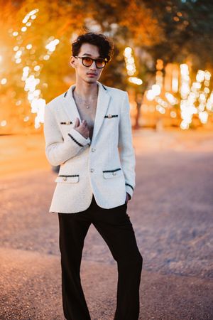 Stylish portrait against orange bokeh lights featuring a white blazer, black pants and glasses in an evening setting.
