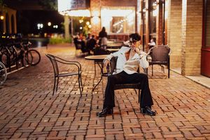 Someone relaxes on an outdoor metal chair at night along a brick sidewalk by a storefront with bicycles parked nearby.