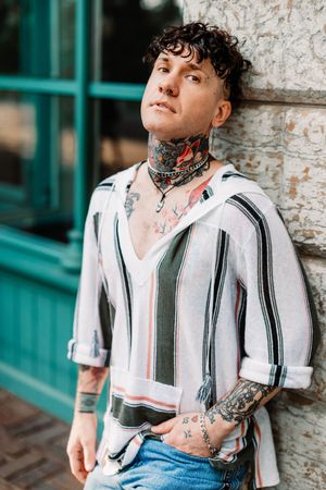 A person in a striped button-down shirt with neck and arm tattoos leans against a brick wall by a green doorframe.