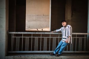 A lone person sits on a chair in a dimly lit industrial corridor with concrete walls and metal railings.
