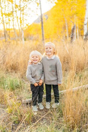 Two children in matching gray sweaters stand together in tall autumn grass in Snowbird, Utah.
