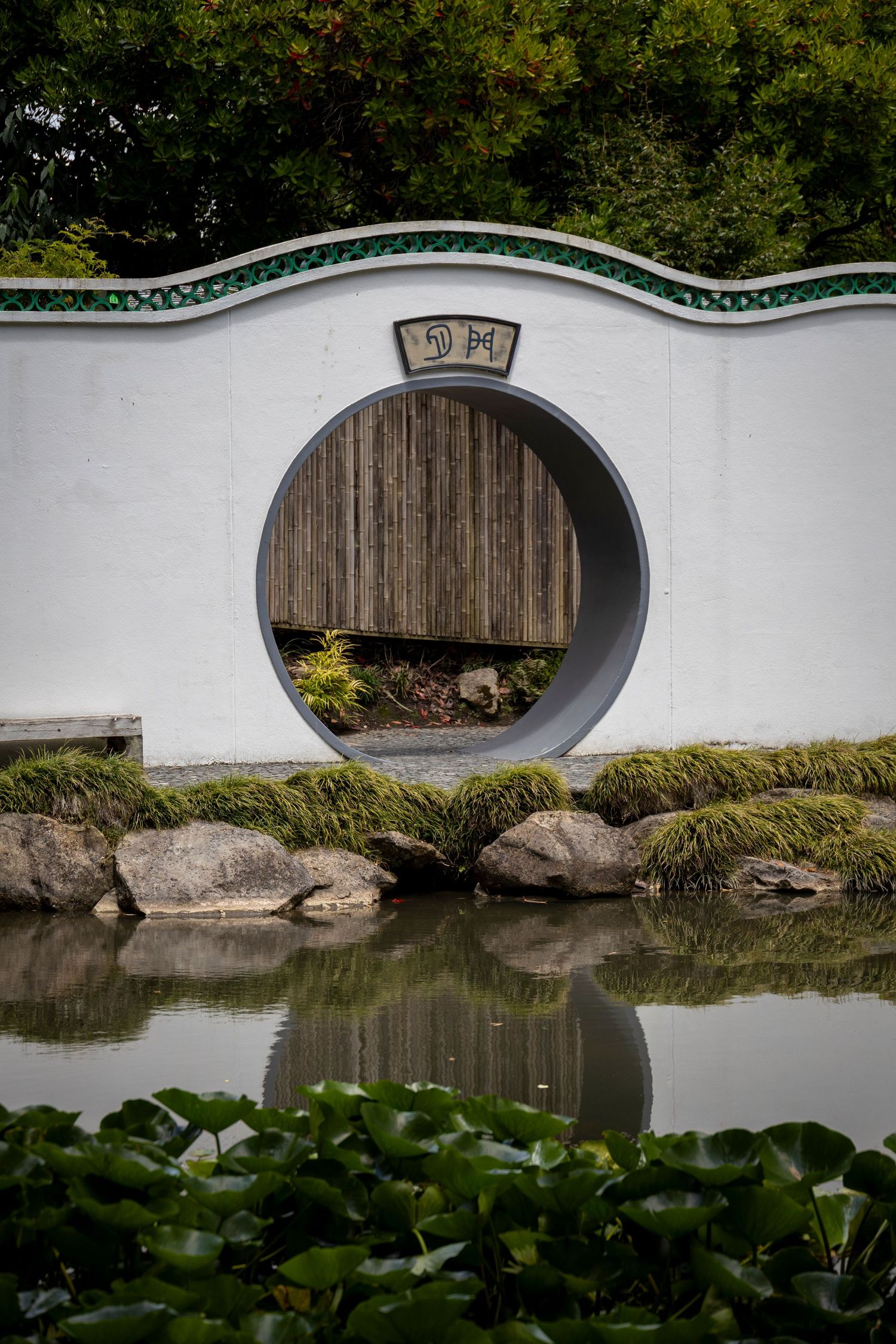 White circular moon gate in a traditional garden wall, reflected in a calm pond with rocks, greenery, and wooden fencing beyond.