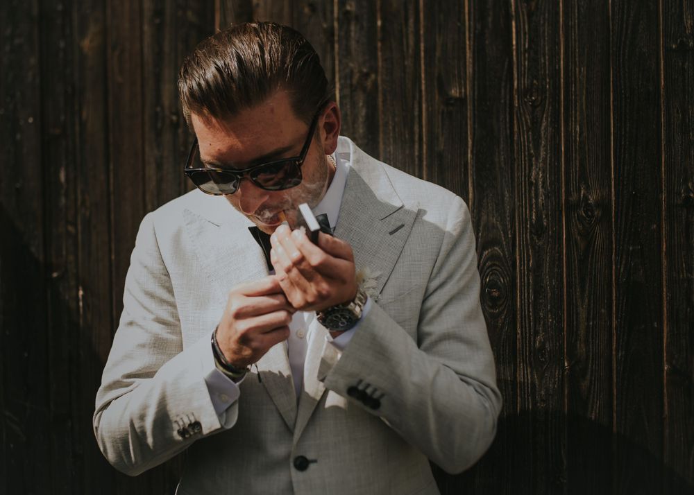 Someone in a light gray suit and sunglasses lights a cigarette against a dark wooden wall background on Fire Island, NY