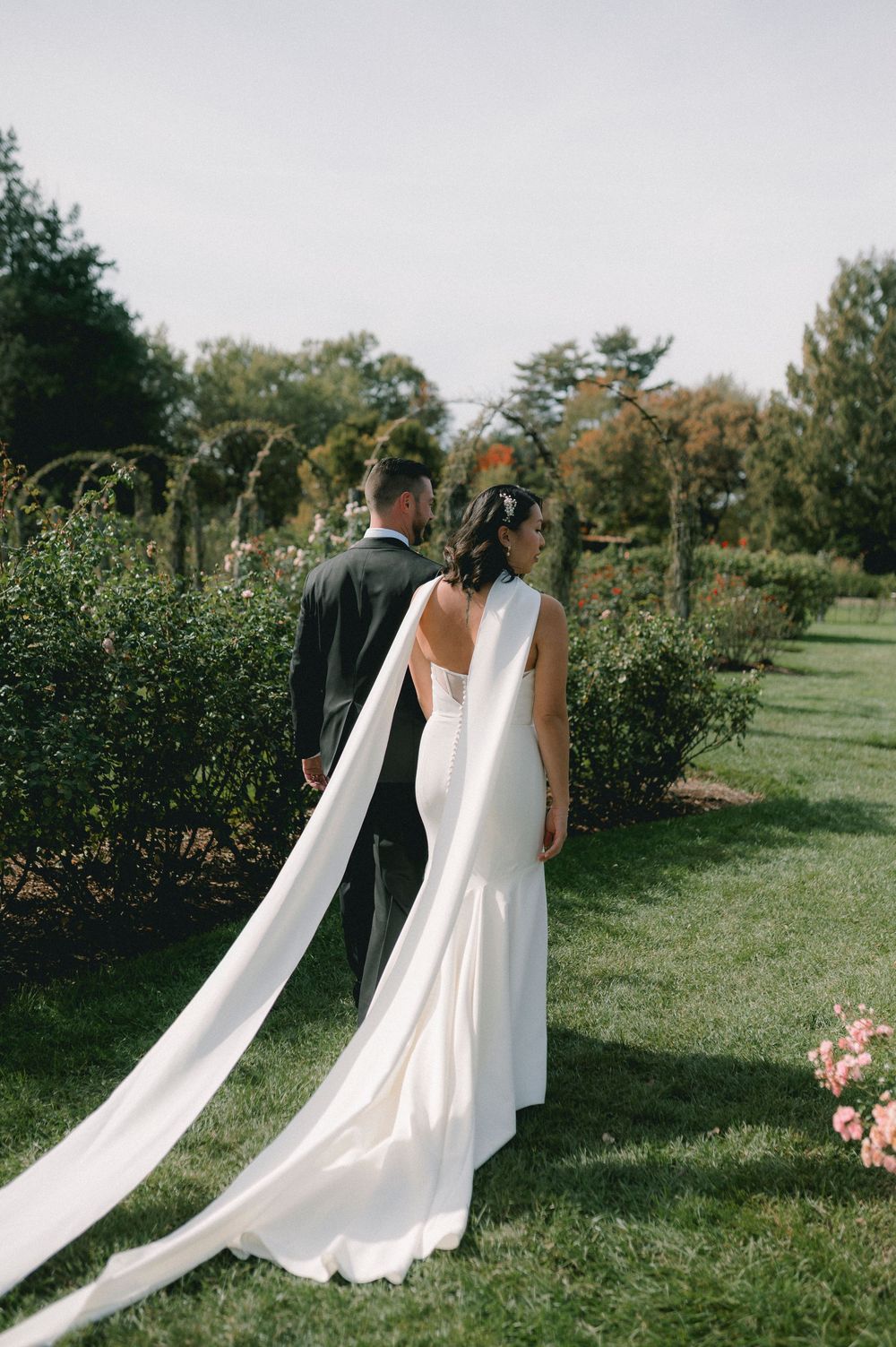 A couple in wedding attire walks through a scenic orchard garden with trailing white dress fabric flowing behind them.