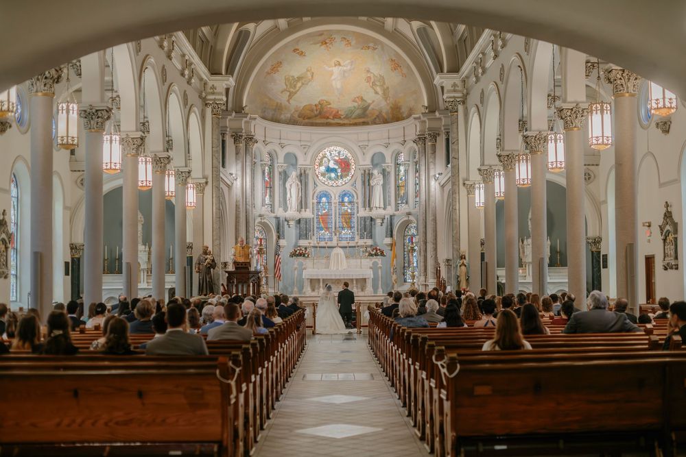 A traditional Catholic church interior with wooden pews, marble columns, and ornate altar during a wedding ceremony.
