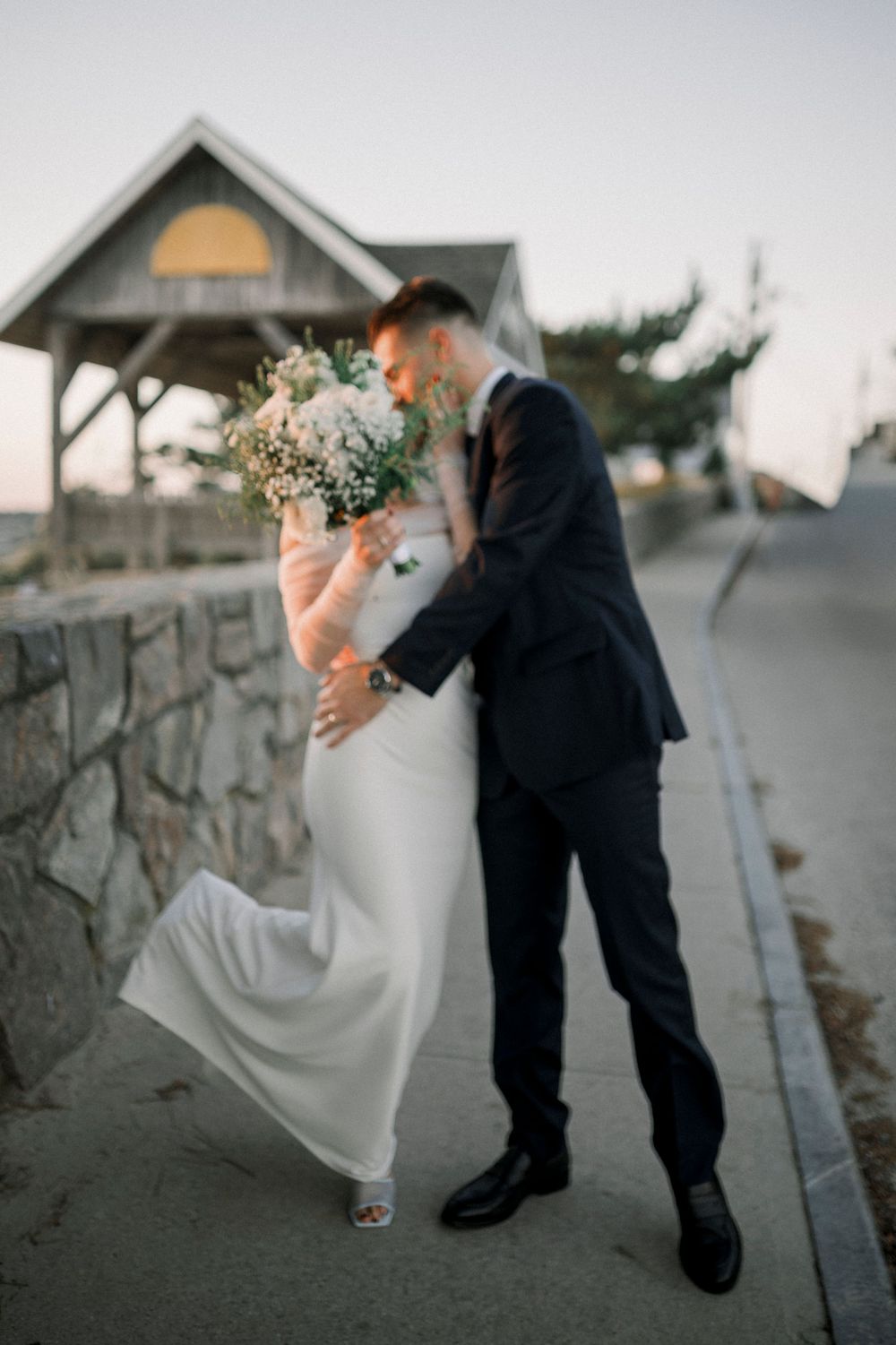 A romantic wedding photo of a couple embracing on a stone path at sunset with the bride holding a white bouquet.