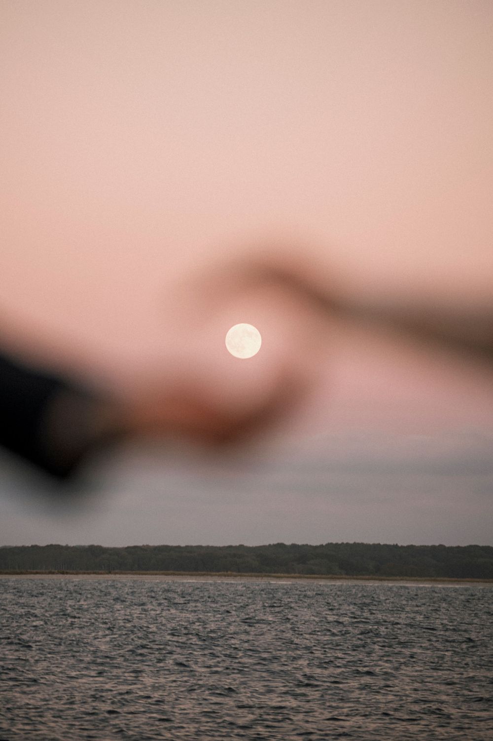 A bright full moon glows in a dusty pink sky over rippling water with a distant shoreline at dusk.