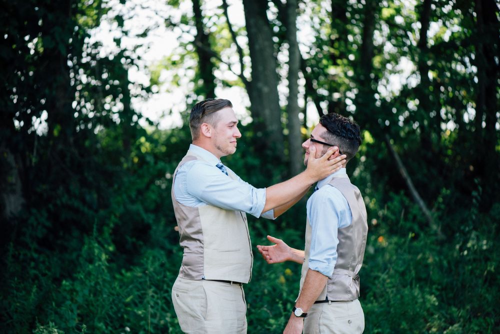 Two people in light colored formal attire share an emotional embrace in a wooded outdoor setting.