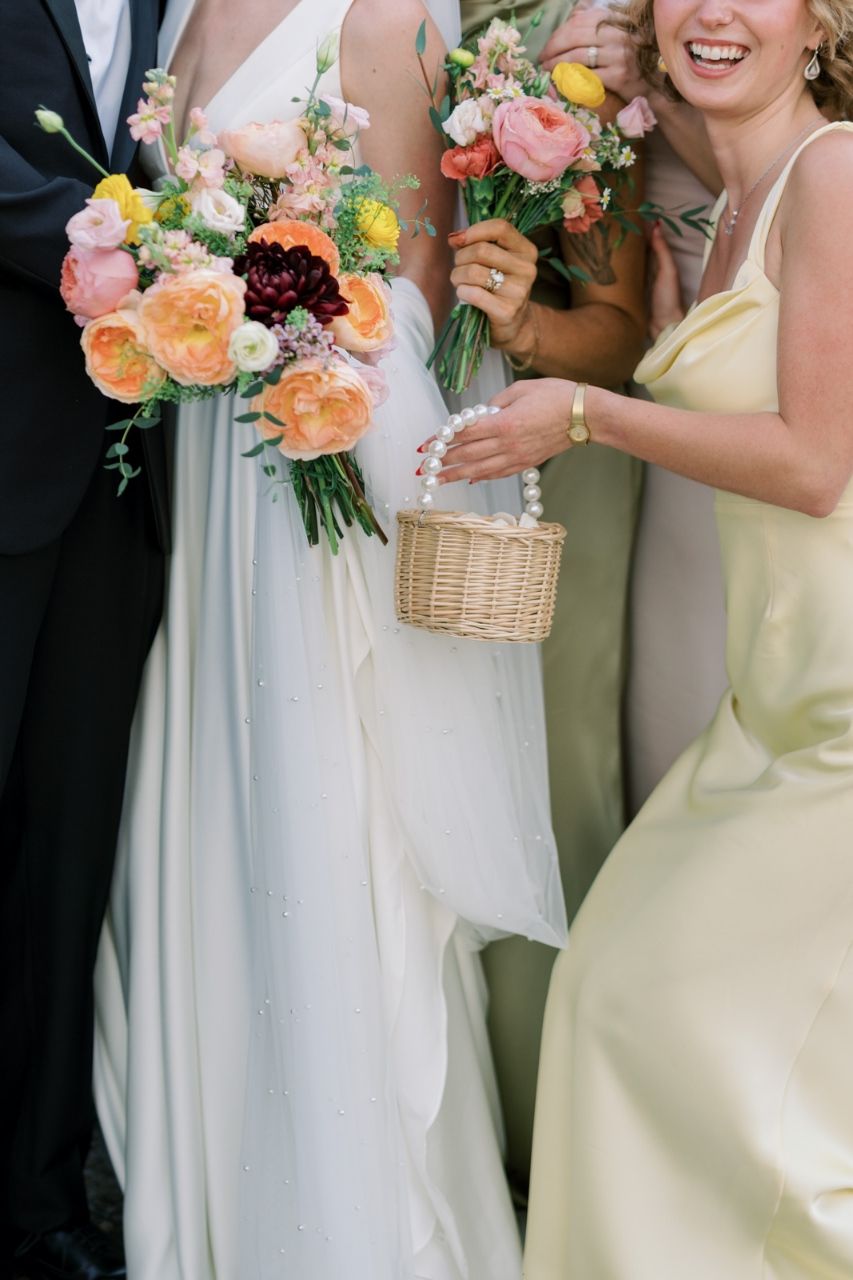 Wedding party members in pastel yellow dresses hold beautiful peach and coral floral bouquets against a light backdrop.