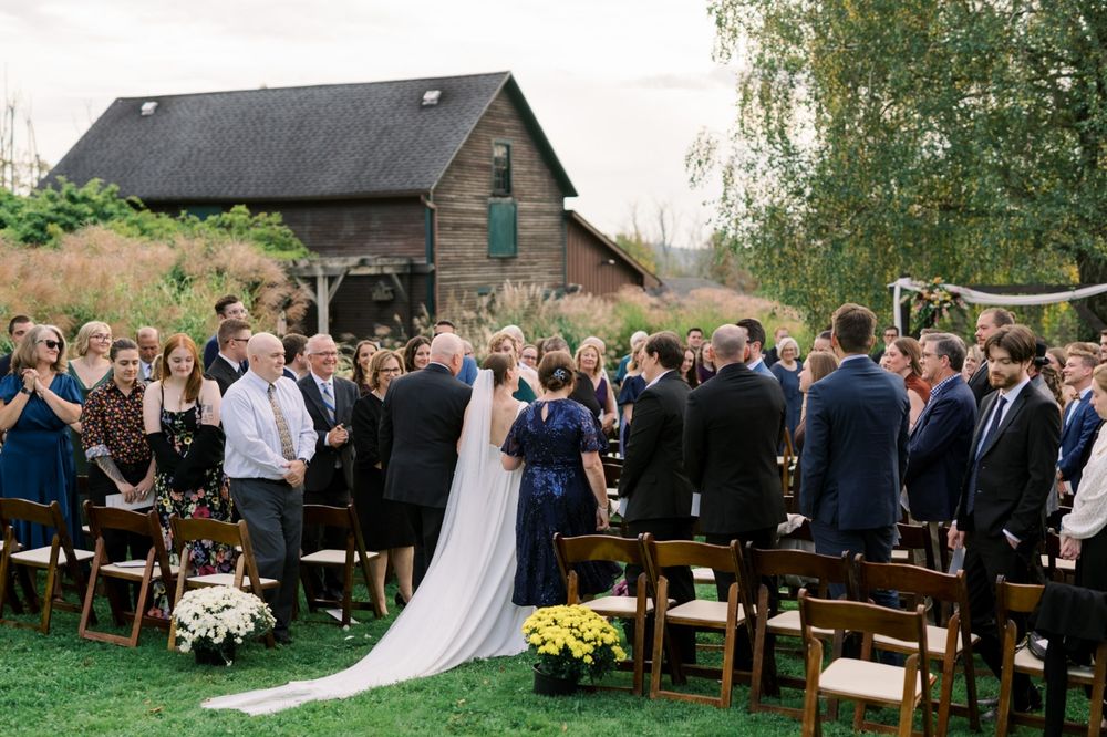 Wedding guests gather outdoors at a rustic venue with wooden benches and natural greenery for a countryside ceremony.