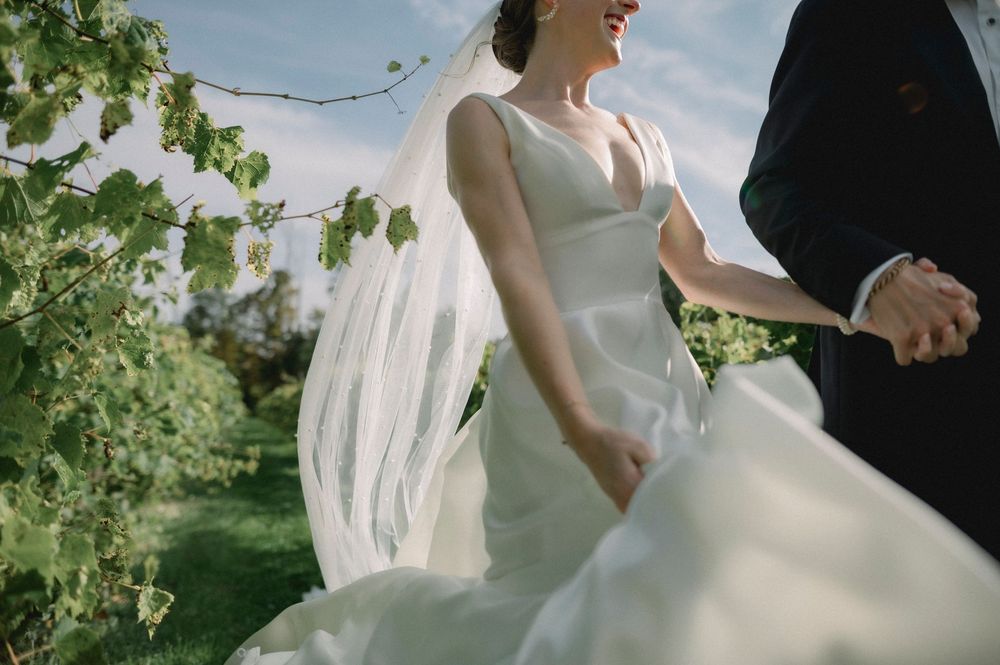 A bride and groom hold hands in a lush garden setting with sunlight filtering through green foliage.