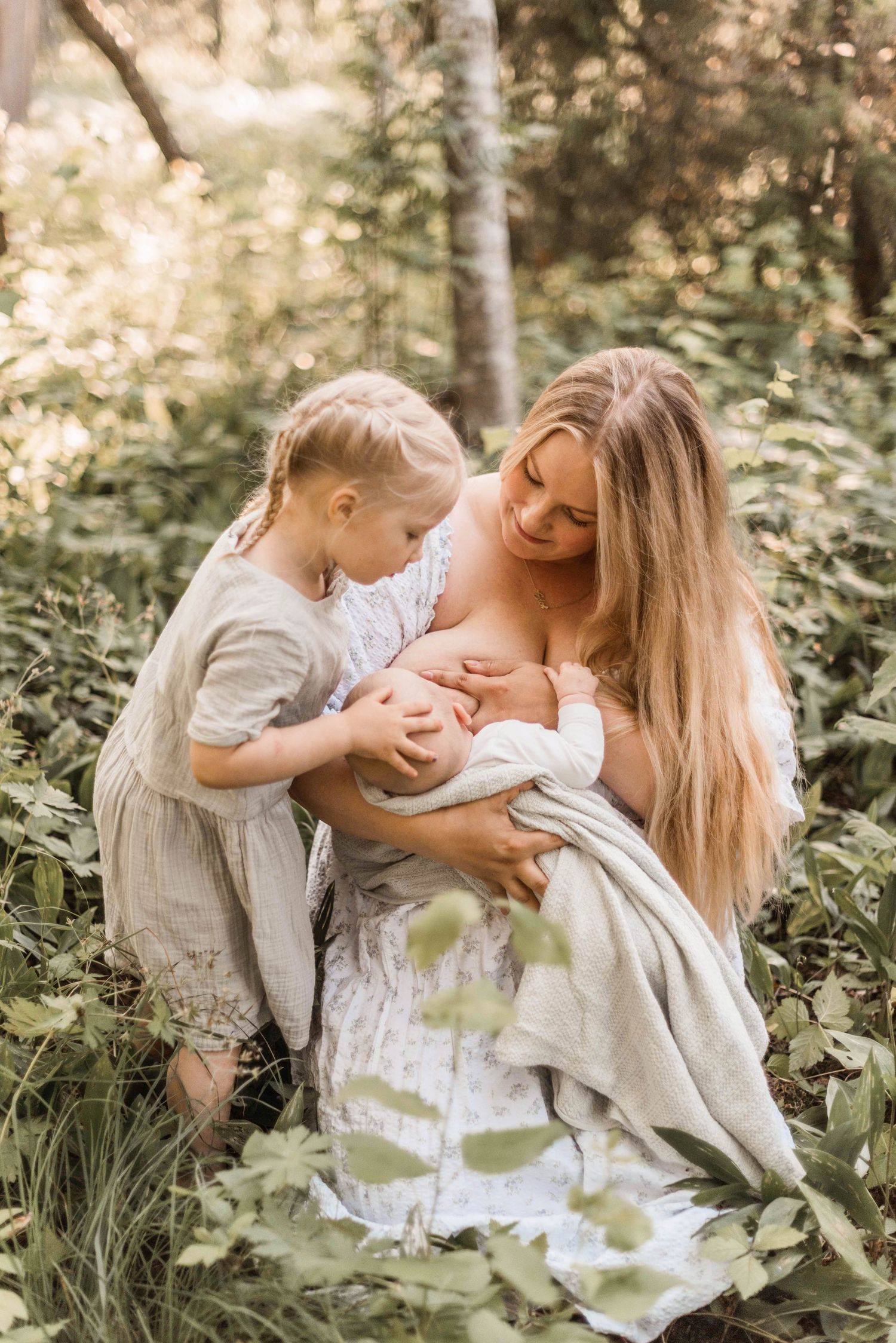 A tender family moment in a sunlit forest clearing as a mother nurses her infant while another child looks on lovingly.