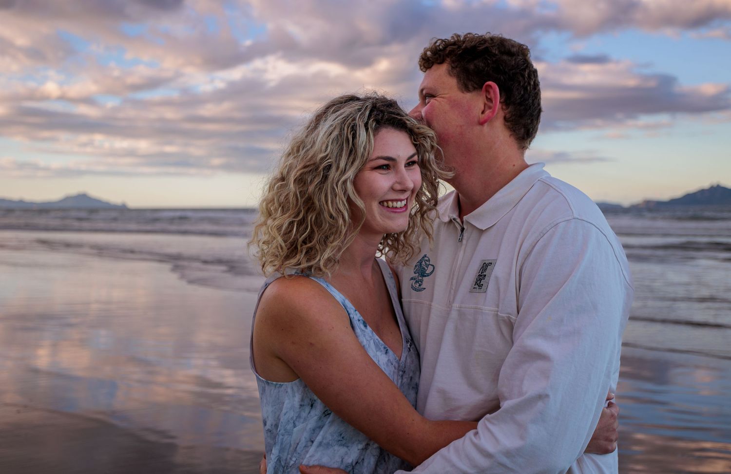 Couple embracing and smiling on beach at sunset with colorful pink and purple sky.