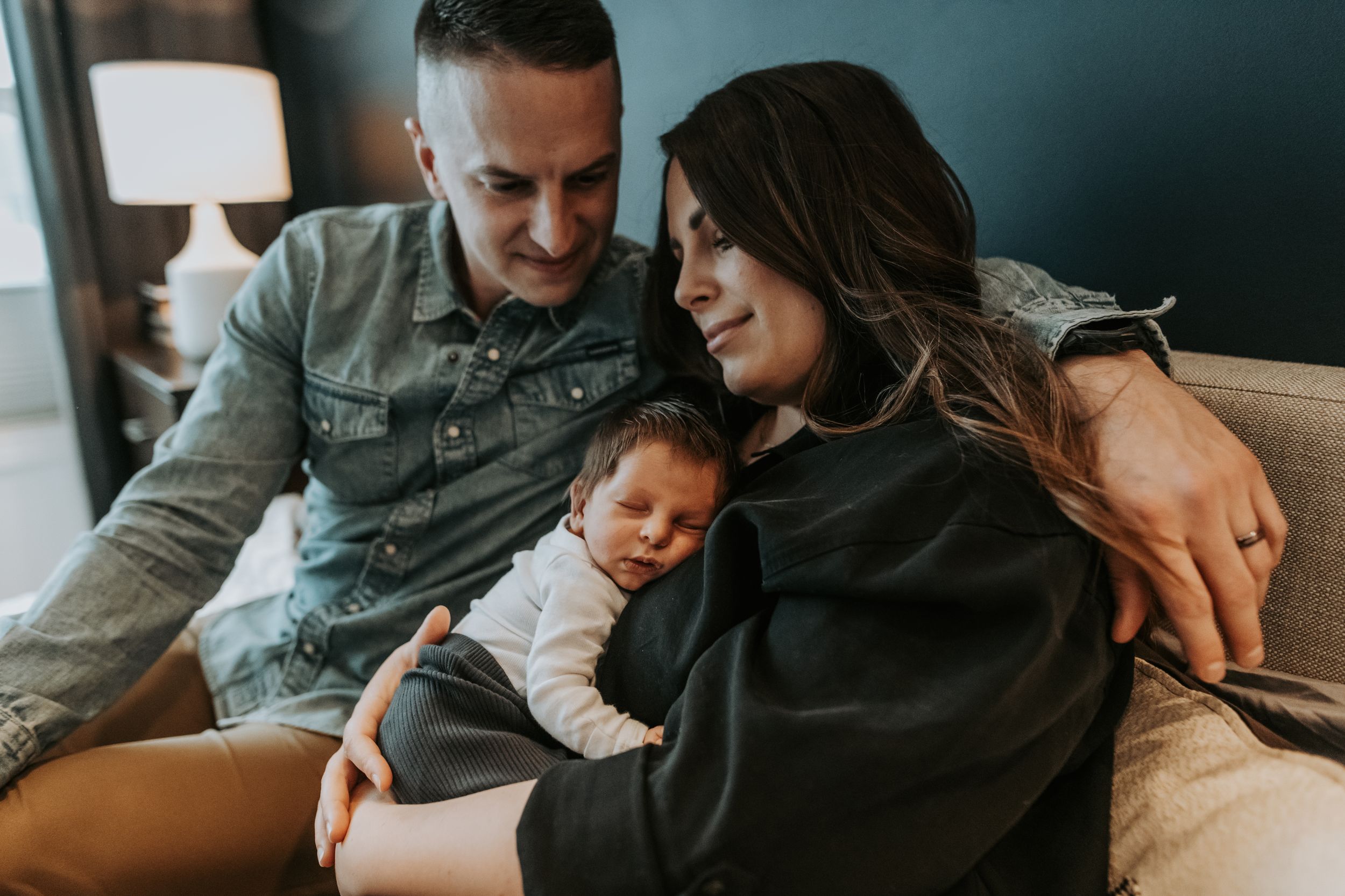 Happy parents sitting on couch together lovingly holding their sleeping newborn baby at home.