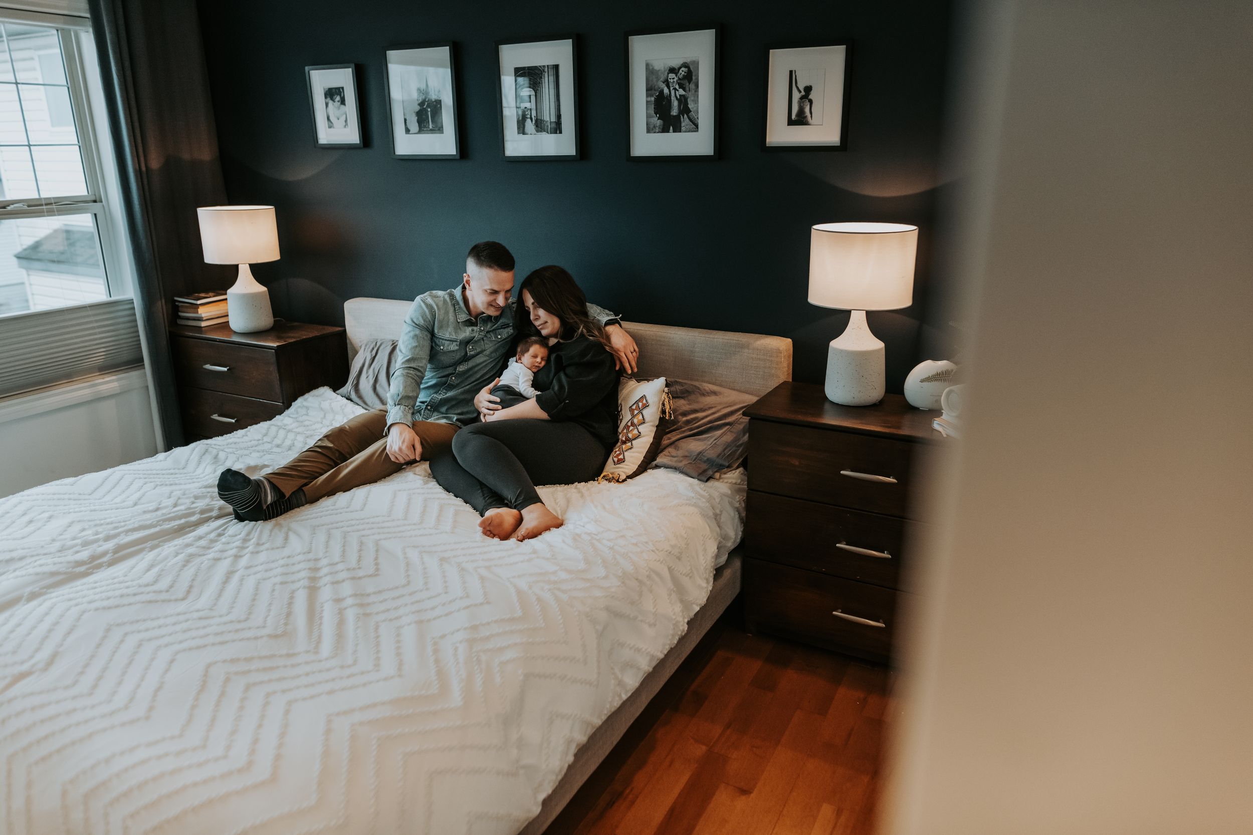 Couple relaxing on bed reading together with newborn baby in cozy dark-walled bedroom.