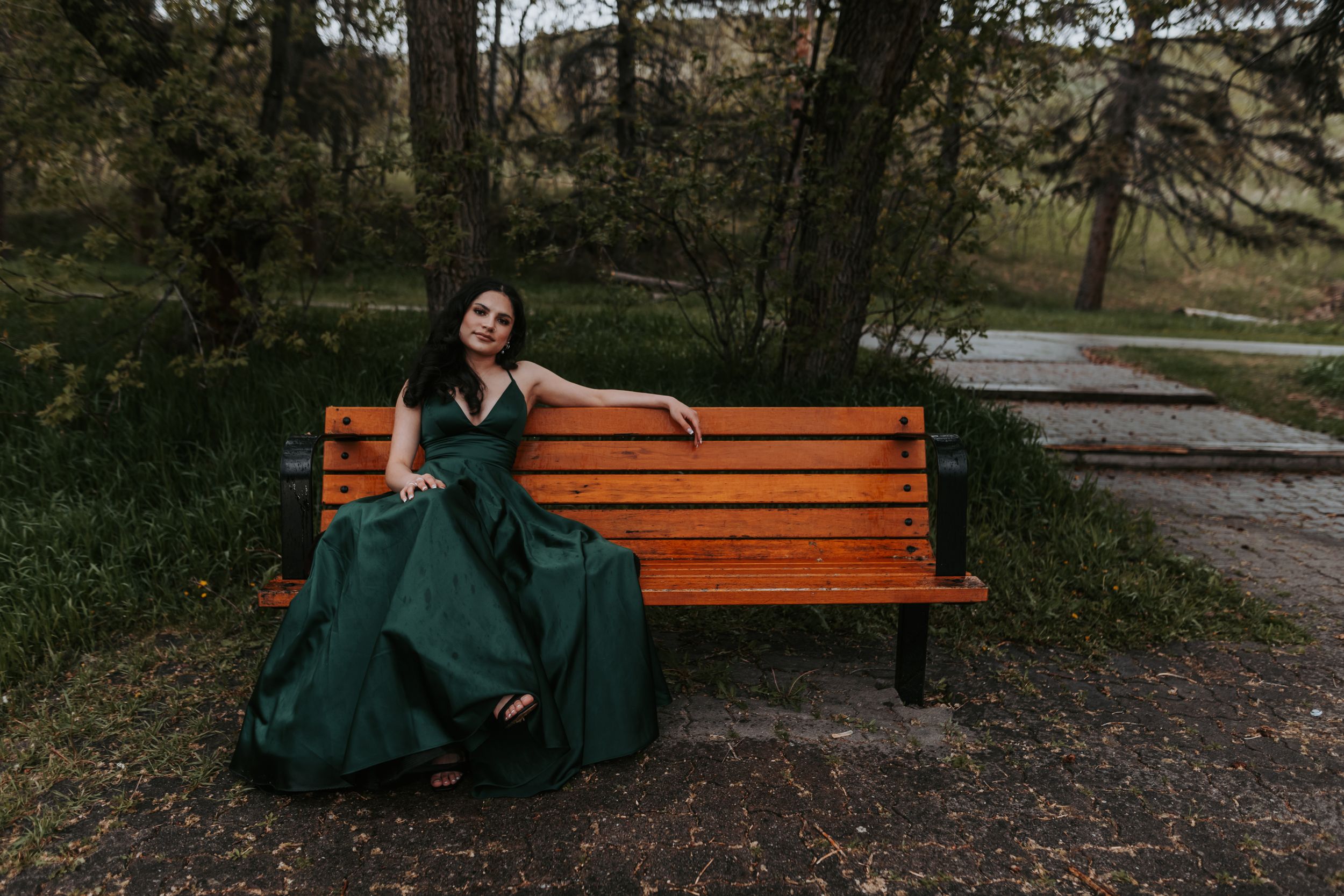 Woman in elegant green ball gown posing gracefully on wooden park bench surrounded by trees.
