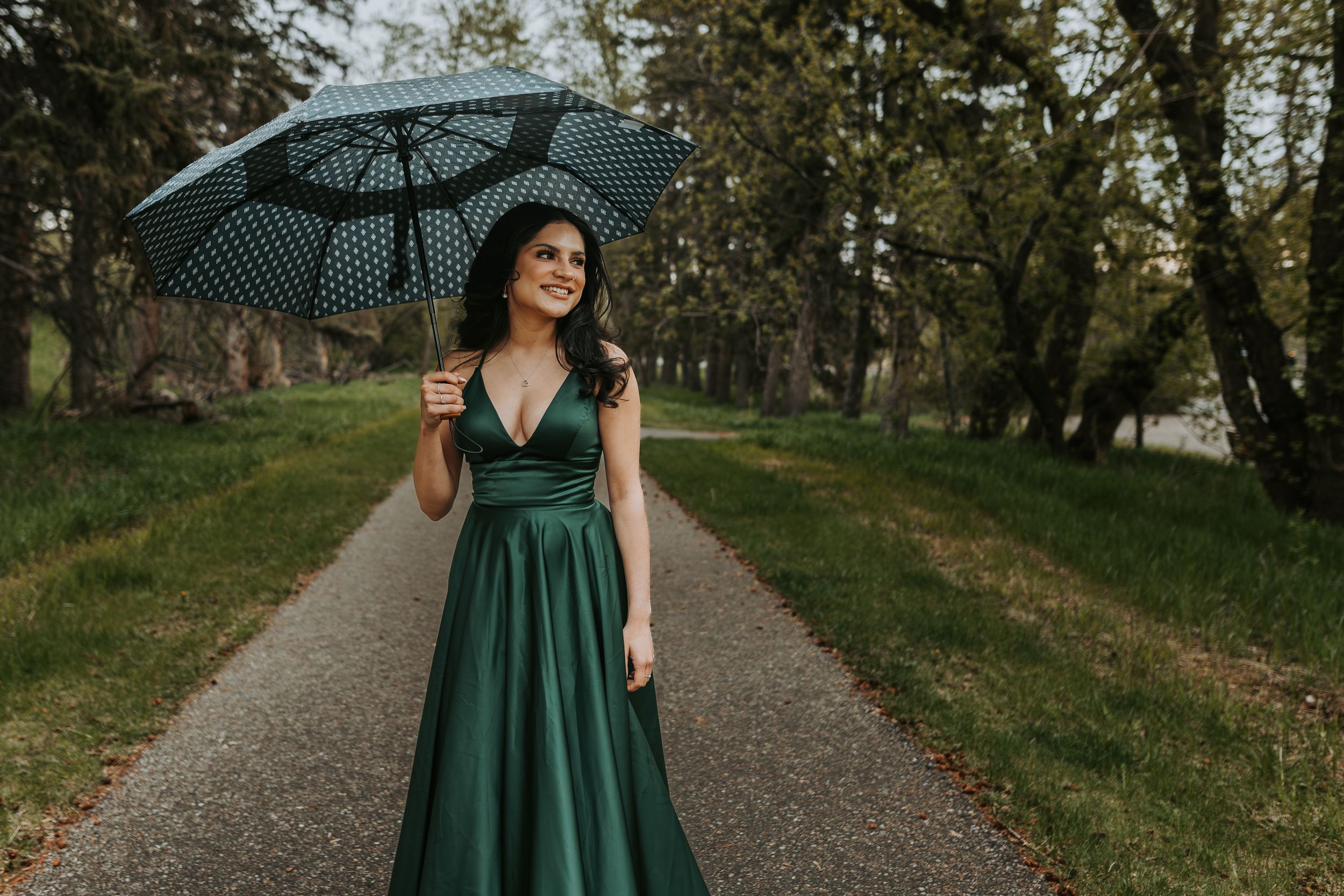 Smiling woman in green satin gown holding polka dot umbrella on tree-lined garden pathway.