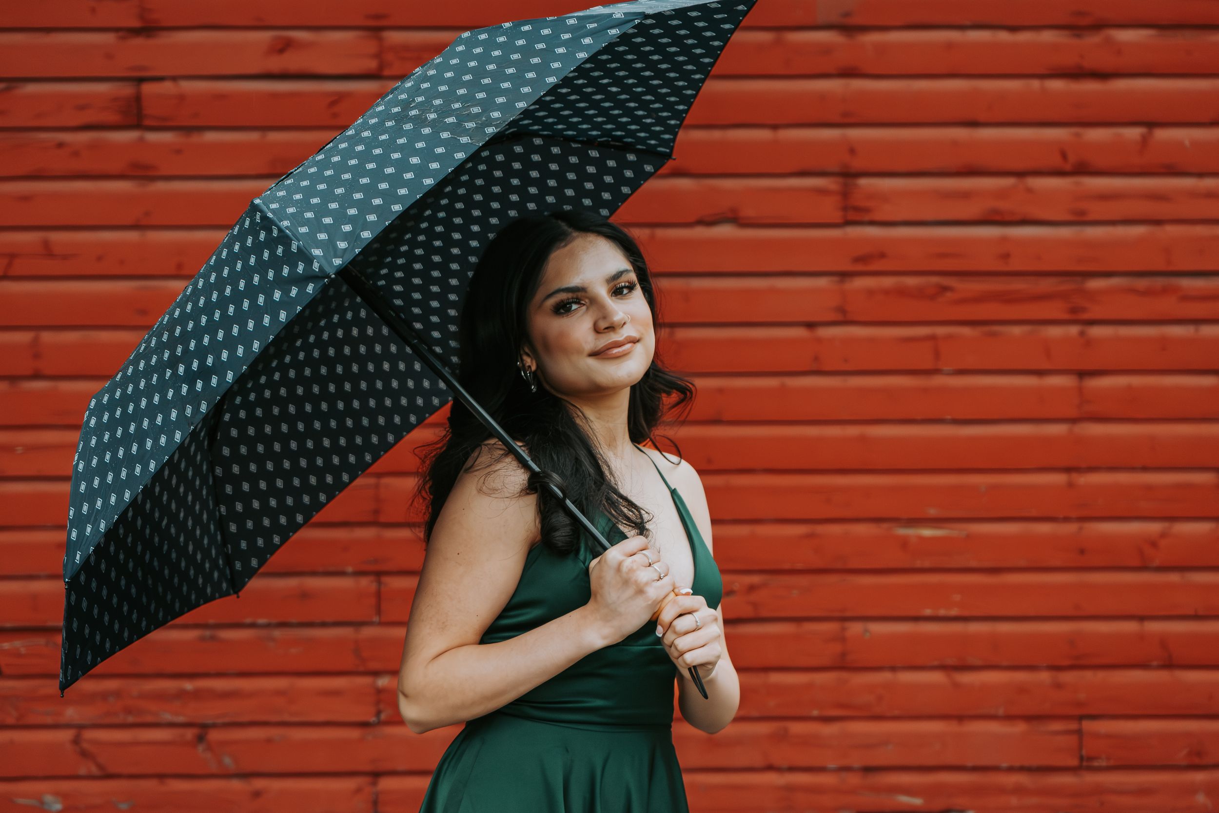 Beautiful woman in green dress holding polka dot umbrella against vibrant red brick wall.