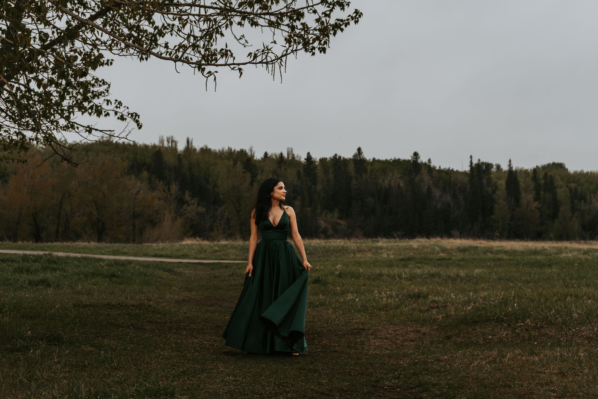 Woman in flowing dark green gown walking through open meadow with forest backdrop on overcast day.