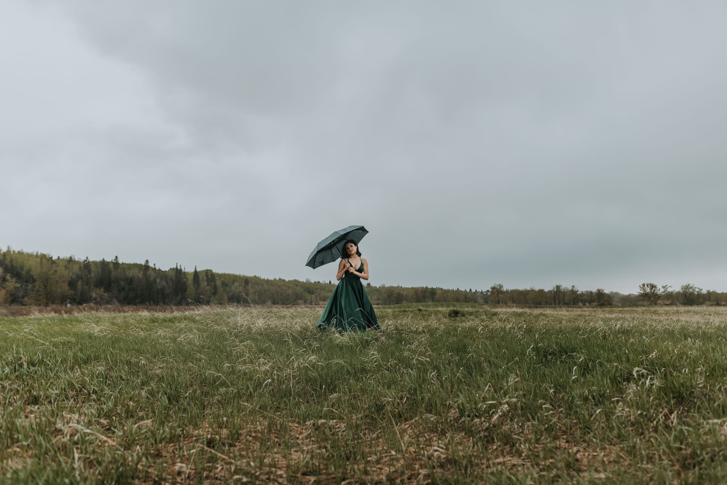 Woman in green ball gown carrying umbrella walking through vast grassy field under dramatic cloudy sky.