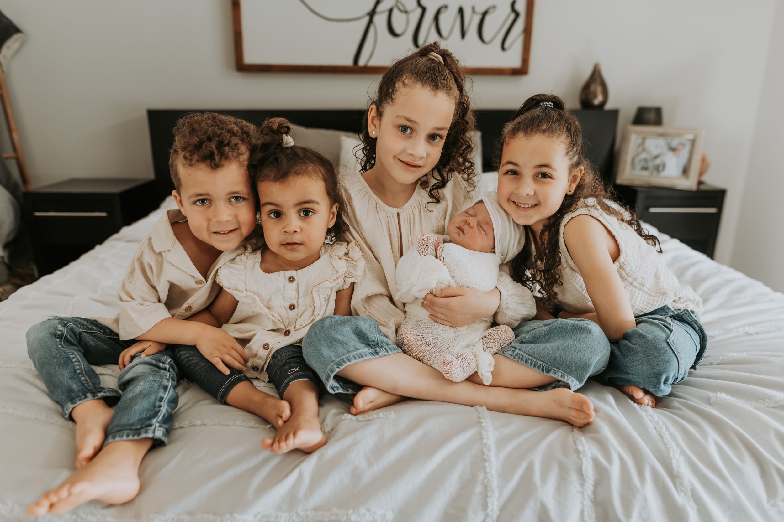 Four young siblings sitting together on white bed, oldest holding newborn baby, smiling warmly.